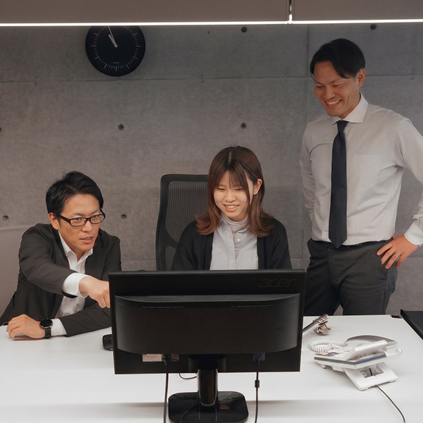 PCモニターを囲み、指を指して熱心にアイデアを共有する男性と、笑顔で見守るメンバー。コンクリート打ち放しのモダンなオフィスで、活発な議論から新しい価値を生み出す様子